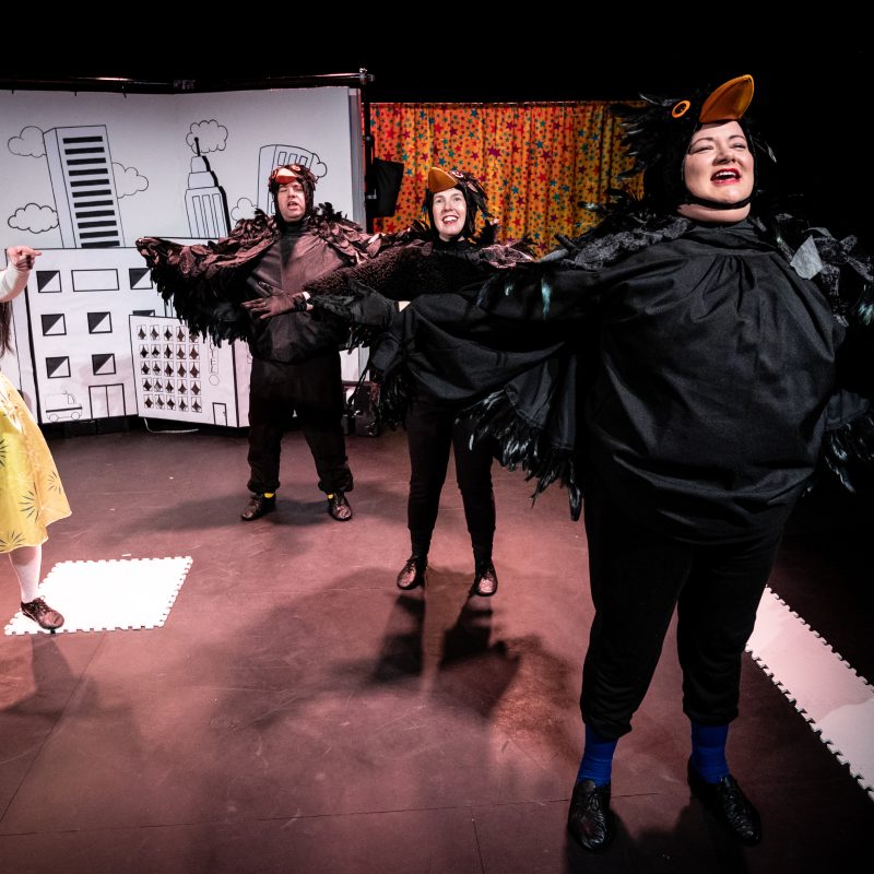 Three Actors stand in a line towards the front of the stage. They wear blackbird costumes with feather wings and hats with orange beaks. To their right in the centre of the stage stands Jasmin, she wears a yellow dress with long white sleeves and white socks with a blue thunderbolt across her chest and holds her cane pointing ahead of them.