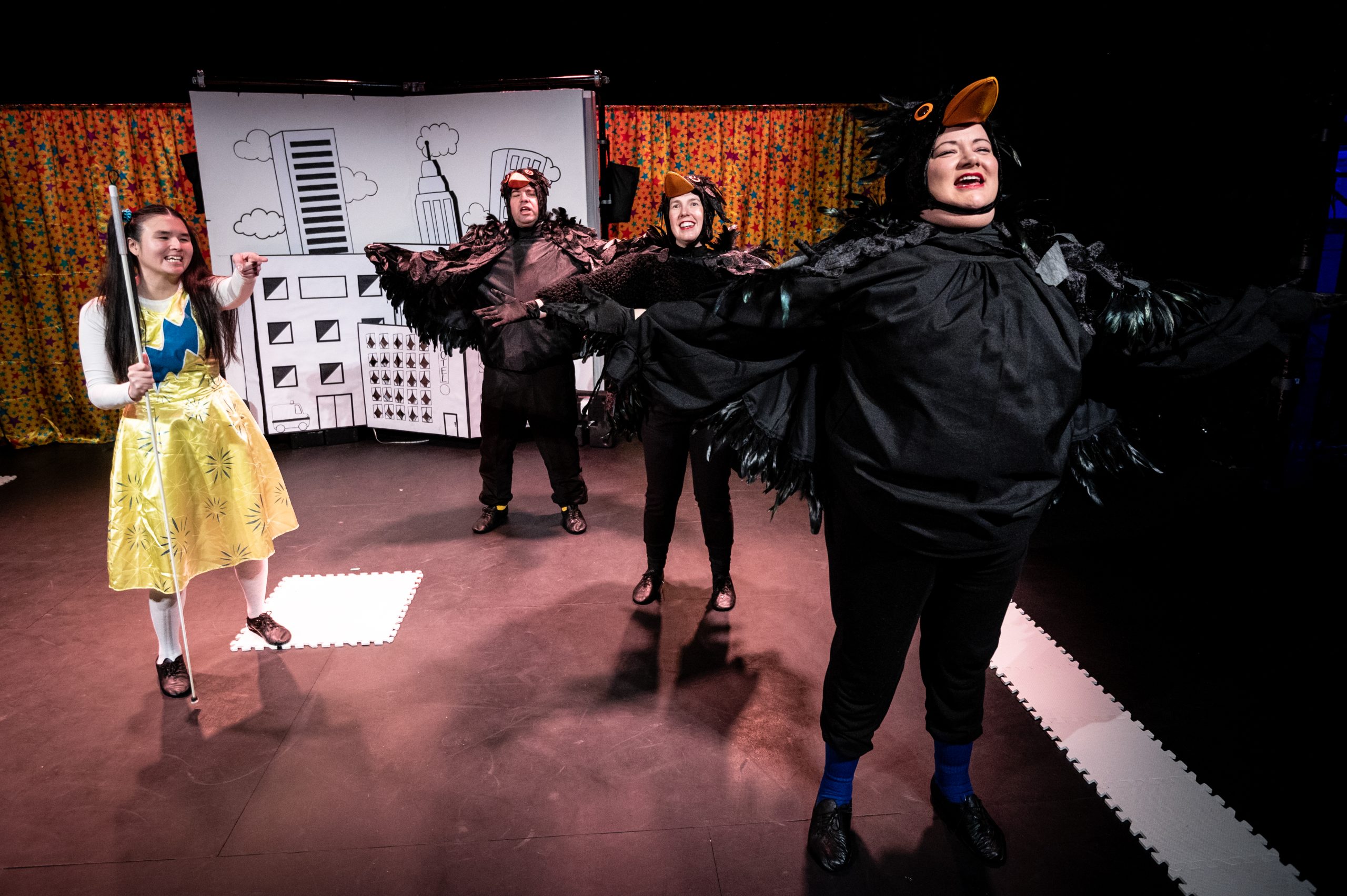 Three Actors stand in a line towards the front of the stage. They wear blackbird costumes with feather wings and hats with orange beaks. To their right in the centre of the stage stands Jasmin, she wears a yellow dress with long white sleeves and white socks with a blue thunderbolt across her chest and holds her cane pointing ahead of them.