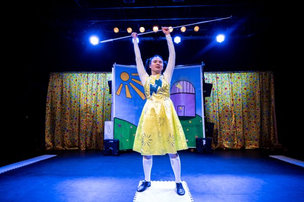 Jasmin, a Chinese actor in her mid-20s stands onstage wearing a yellow dress with a blue lightning bolt across the chest with long white sleeves and socks. She holds a cane triumphantly above her head in both hands.