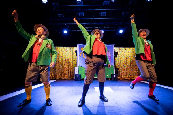 Steve, a white male actor, Chloe, a mixed-heritage female actor and Chris, a white male actor stand in a line at the front of the stage with their right hands raised. They all wear boater hats, green blazers with red waistcoats underneath, grey shorts and long colourful socks.