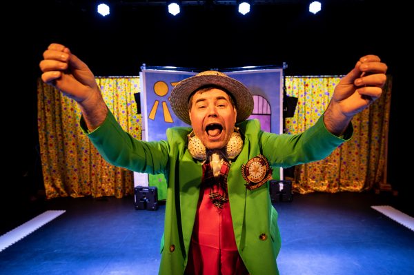 Steve, a white actor wearing a boater hats, green blazer with a red waistcoat underneath, and a pair of golden headphones around his neck looks directly into the camera with his arms outstretched and his mouth open wide as if shouting.