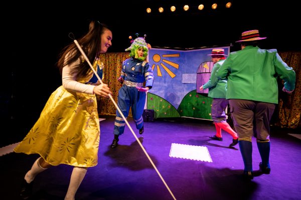 Four actors are running in a circle around the centre of the stage, engaged in a panto chase scene. Two of them wear blazers, boater hats and long socks, one a yellow dress and one a blue boiler suit with a bright green wig.