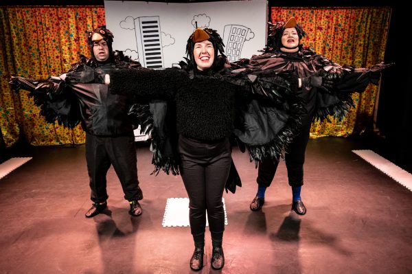 Actors Steve, Angela and Chloe stand in a triangular formation in the centre of the stage with their arms outstretched as though flying. All three of them are wearing blackbird costumes with feathery wings and hats with orange beaks.