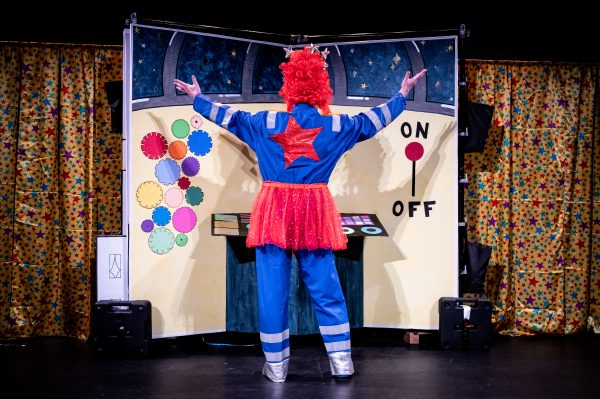An actor wearing a blue boiler suit with a bright red star on the back, a tall red wig and a red tutu has his back to the camera and his arms raised towards the colourful yellow observatory set.