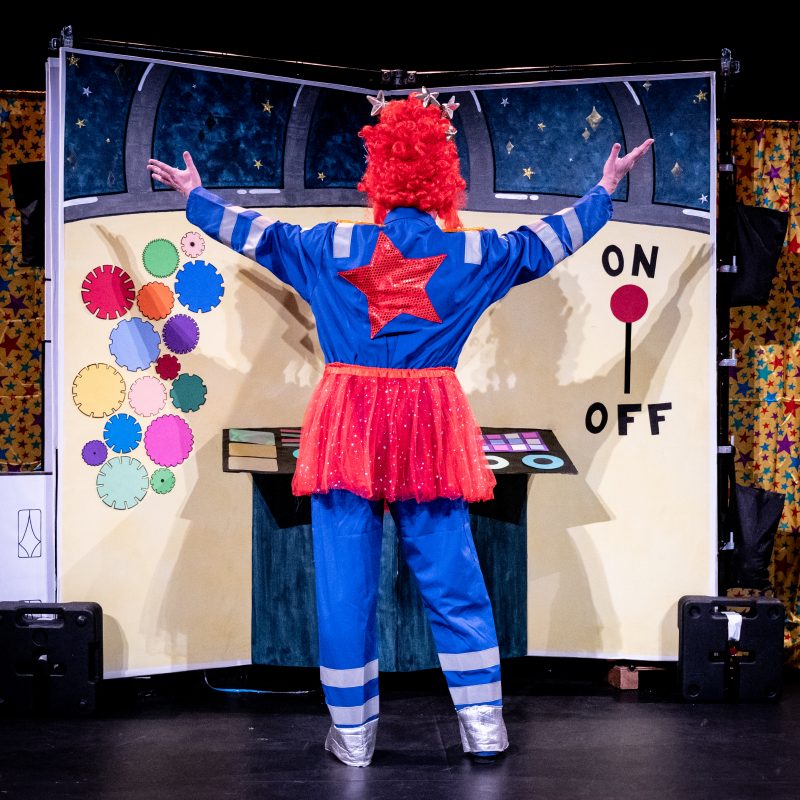 An actor wearing a blue boiler suit with a bright red star on the back, a tall red wig and a red tutu has his back to the camera and his arms raised towards the colourful yellow observatory set.