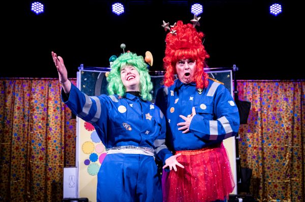 Angela, a white female actor wearing a blue boiler suit and bright green wig decorated with little planets stands smiling with her arms outstretched as though in song. Next to her stands Chris, a white actor wearing a blue boiler suit with a bright pink tutu and tall wig of the same colour decorated with stars. he has his hand on his torso and pulls an expression as though singing a low operatic note. They stand in front of the Super Power Panto comic book observatory set, either side of which is a yellow curtain covered in red and green stars.