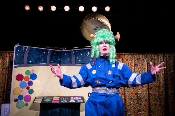 Angela, a white female actor wearing a blue boiler suit and bright green wig decorated with little planets stands with her hands outstretched expectantly while behind her a large golden beach ball is flying over the observatory set in her direction. 