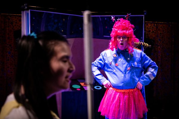 Out of focus close in the foreground is Jasmin, a Chinese female actor with pig tails holding a white cane. In focus in the background is Chris, a white actor wearing a blue boiler suit with a bright pink tutu and tall wig of the same colour. He pulls a shocked expression looking towards Jasmin.