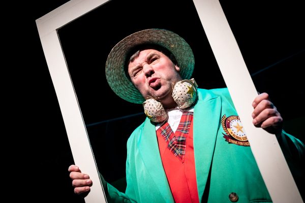 White actor Steven George stands with a scrunched up facial expression holding a white frame around himself. He wears a boater hat, green blazer with a red waistcoat underneath and a pair of golden headphones around his neck.