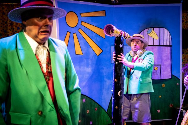 White actor Chris Biddle pulls a worried expression while white actor Steven George aims a red and gold telescope on a stand decorated with stars and moons at him. Both actors wear boater hats, green blazers with red waistcoats underneath, grey shorts and long socks. Steve is also wearing a pair of golden headphones. They are in front of a comic book set of the exterior of an observatory.