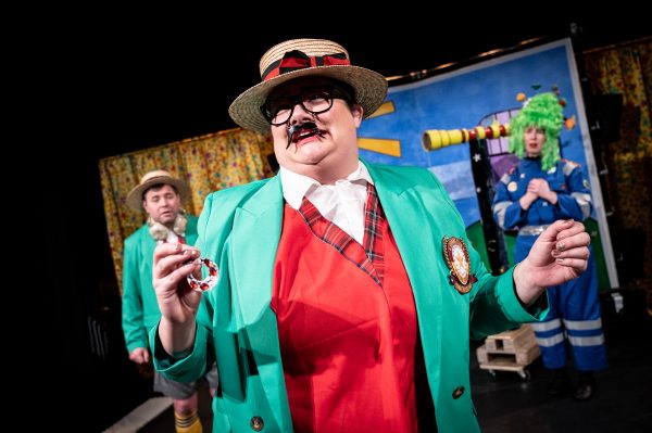 Mixed heritage actor Chloe Driver stands in the middle of the stage wearing a boater hat, green brazer, red waistcoat, white shirt, glasses and false moustache. Behind her is one male actor, dressed the same as her, and one female actor wearing a blue boiler suit and green wig. They are stood in front of a set of starry yellow curtains and the Super Power Panto outdoor observatory set.