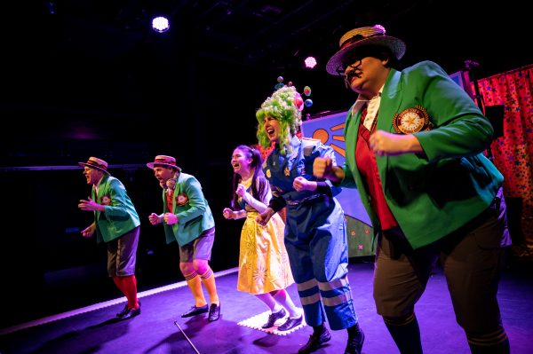 All five super power panto performers onstage in a line in the middle of the show's final number. They're all smiling with their fists clenched in front of them. Three of them wear blazers, boater hats and long socks, one a yellow dress and one a blue boiler suit with a bright green wig.