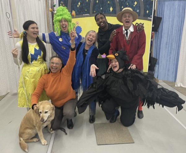 A group photo of the cast of Super Power Panto in costume looking towards the camera and smiling with their arms outstretched. Also in the group are directors Shannon and Maria (with her guide dog Bella) and actor Kiell Smith-Bynoe.
