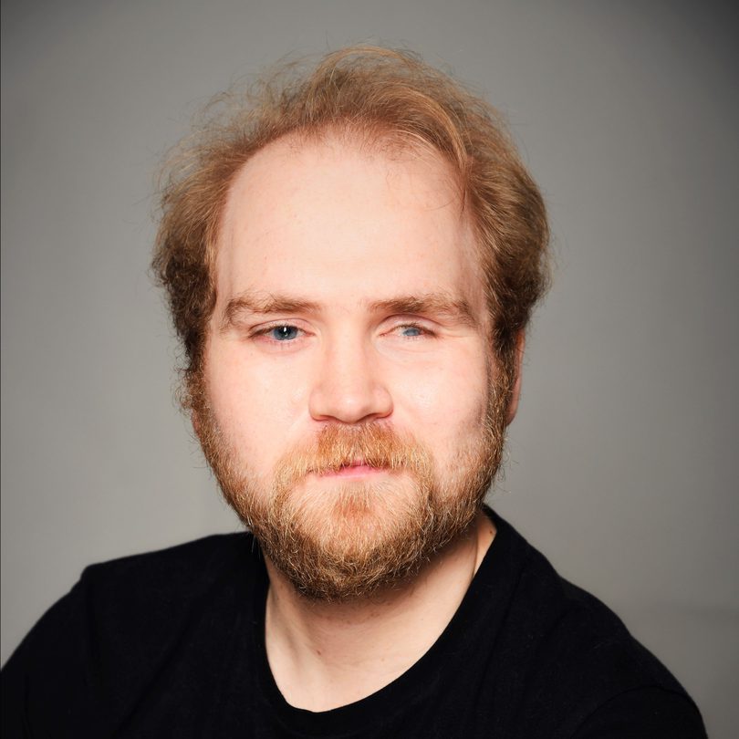 This image is a headshot of Extant's new Trainee Artistic Director Ben Wilson. He is a white man with short blonde hair, blue eyes, and a beard. He wears a black t-shirt.