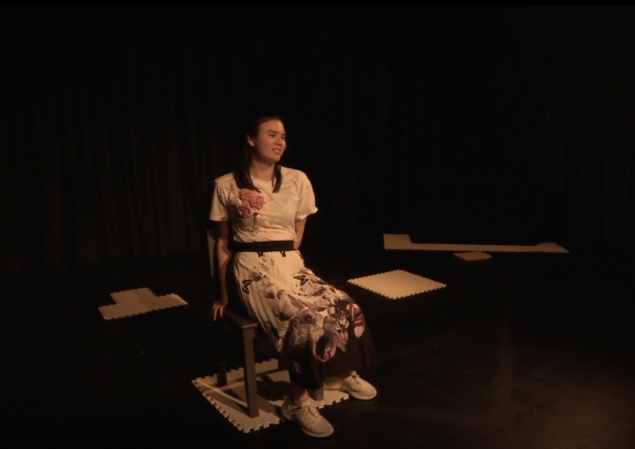 Jasmin Thien sits on a wooden chair in a black box theatre. Jasmin is a young Chinese-British woman with long dark hair pinned back off her face. She wears a white top and skirt decorated with floral motifs, and white trainers. The floor under the chair and around the stage is marked with white tactile flooring tiles.