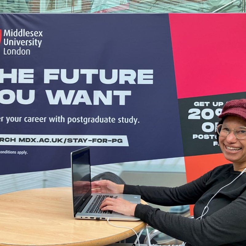The image shows Maria sitting at a round table. To her right-hand side is a large pink and blue banner. In the top left-hand corner is the coat of arms for Middlesex university. Underneath this, in large capital letters, are the words, ‘ The Future You Want - Master your career with postgraduate study’. The photo is taken opposite Maria. We see her laptop which is open on the table, with one of her hands on the keys. Her cane sits unfolded on her lap. She wears a black long-sleeved T-shirt, a burgundy peaked cap and metal framed glasses. She looks into the camera with a big smile on her face.