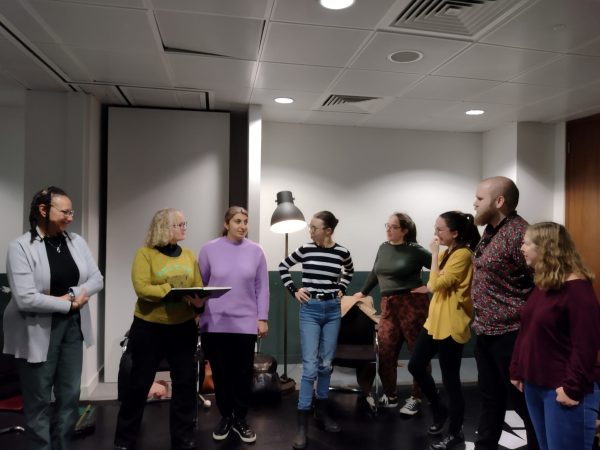 A group of eight people of varying ethnicities and genders are rehearsing and smiling together. They are dressed colourfully and casually in a brightly lit white and green room.