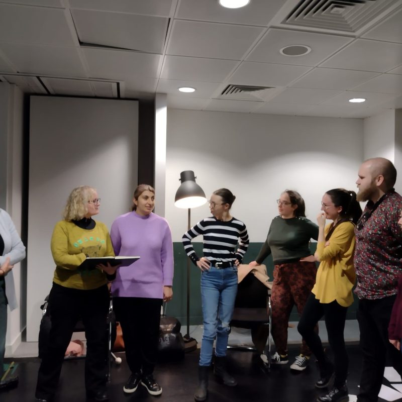 A group of people from Extant Unseen rehearsals standing in a line in a rehearsal room at Theatre Deli. From left to right is Maria Oshodi, Anna Clarkson, Tafsila Khan, Georgie Morrell, Louisa Sanfey, Esther Irving, Ben Wilson and Tam Gilbert.