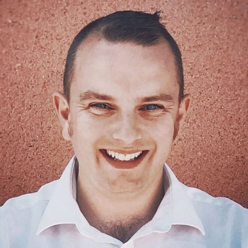 Ian is a white man, in his late 30s, clean shaven, with a small quiff and closely cropped hair. He is smiling and is wearing a white shirt and there is a terracotta wall behind him.