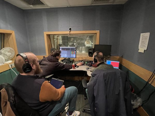 A photo from behind of three white people sitting at a recording studio desk. From the left, Ben is sat back wearing headphones and holding an IPad, Han is leaning over and pressing a button and Ian is listening to the recording.