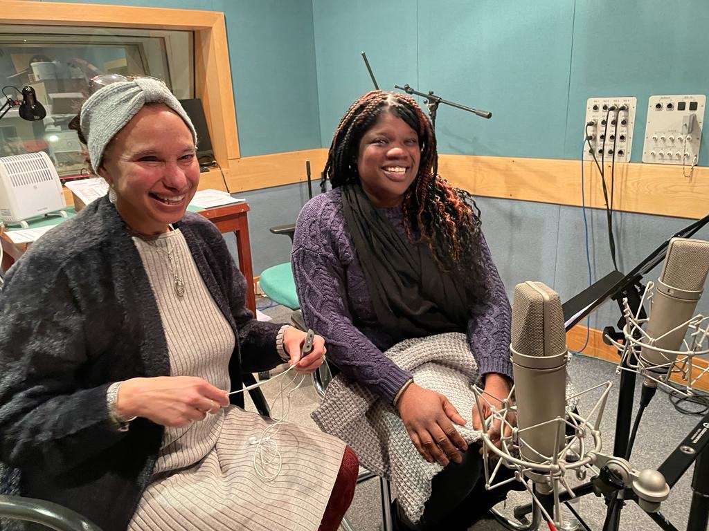 A photo of Maria, a middle aged biracial woman, sitting next to Anita, a young black woman who are together and smiling in a recording studio.