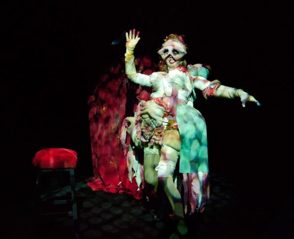 A Caucasian woman stands in front of a red curtain, next to a red stool, with bandages on her arms and legs, and has a fabric vagina pinned to the open skirt of her dress. She also has large fake breasts and exaggerated make-up on a mask that she wears.