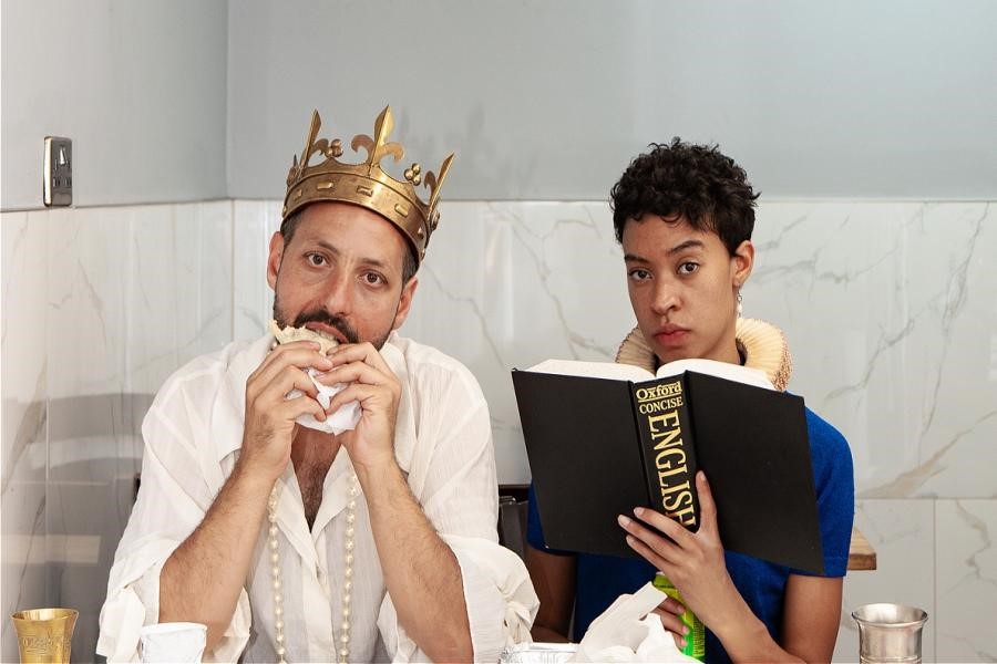 A man is wearing a gold crown and a white shirt next to a woman with a regal collar and a blue shirt. They are eating food in a marbled restaurant. The man is taking a bite of his sandwich and the woman is reading the Oxford English dictionary. 