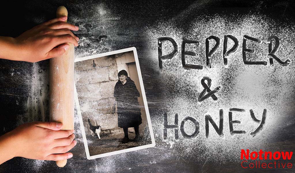 A table covered in flour with a rolling pin rolling over a picture of an old lady dressed in black and a black and white cat. Text is written by hand in to the flour, it reads: Pepper and Honey.