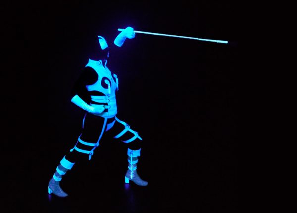 A figure stands in black light with glowing white aspects of costume picking out their fencing pose – side on to the camera, legs wide apart, knees bent, right arm on right hip and left arm above their head pointing forwards holding a glowing white cane thrust forward like a rapier.