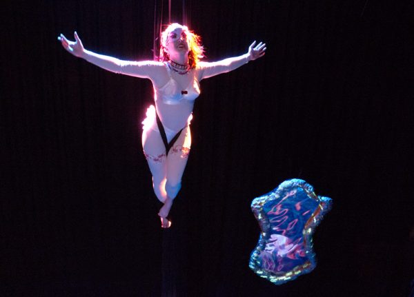 A Caucasian woman is ‘flying’ in mid-air, suspended by her waist. She wears a nude bodysuit and a white pointed bra. She is also wearing an embellished necklace and a half-face mask. Her legs are together and pointed and her arms are flung wide. She is smiling. Beneath her hangs a large glittering fairy tale style mirror.