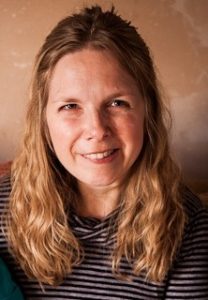 Tam has a broad smile. Her blonde, curly hair is shoulder length, and is she is wearing a predominantly blue and grey striped hooped top.