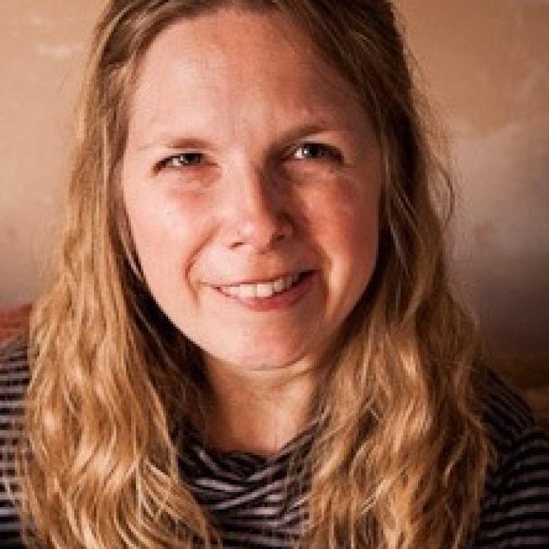Tam has a broad smile. Her blonde, curly hair is shoulder length, and is she is wearing a predominantly blue and grey striped hooped top.
