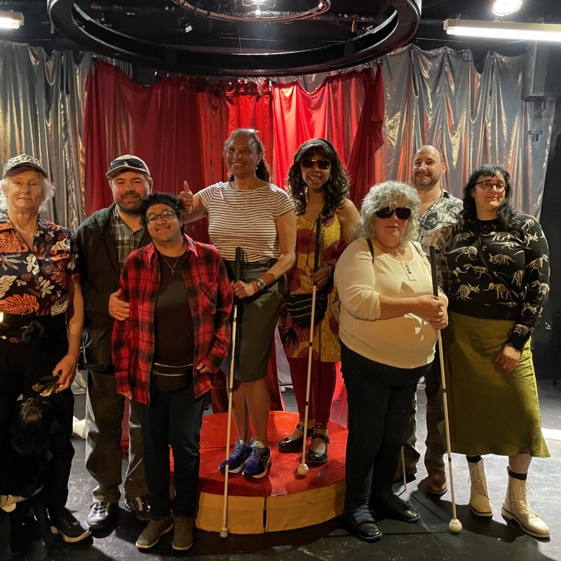 A group of six women and two men are standing on the stage at Camden People's Theatre, posing for a photo and smiling. They are a diverse group of adults, of various ages and ethnicities. Three of them are holding their white canes and there is a guide dog to the left of the photo.