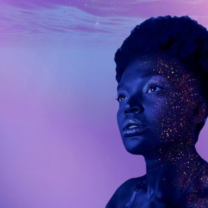 A Black woman’s face, partly covered in sparkling glitter. She intently and blankly gazed outside the frame. The background is filled with gentle water waves in light shades of purple and blue. 