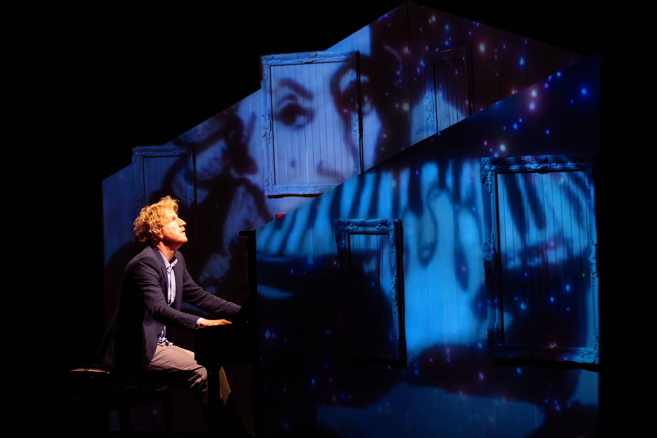 A person sits at a piano onstage, illuminated under blue and white light with projected images of faces and stars on the background wall.