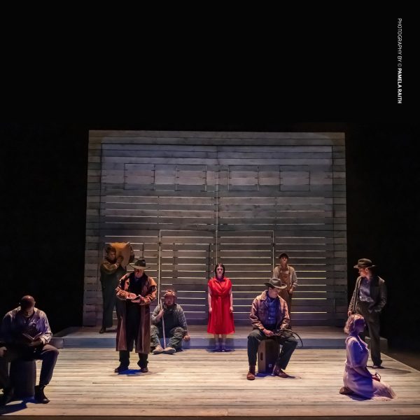 A stage performance of Of Mice and Men features a wooden set with slatted walls, dim lighting, and nine actors in period-appropriate 1930s attire. The central figure, a woman in a striking red dress, stands among male characters dressed as ranch workers and drifters, creating a dramatic tableau. 