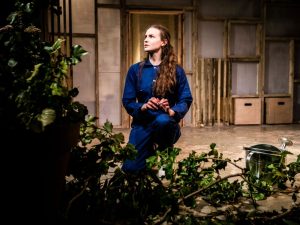 A white woman, with long brown hair, wearing a blue jumpsuit stairs into the distance over a bush. 