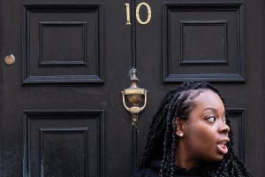 A cropped photo of a black woman's head who looks to her left and stands outside a black door labelled number 10.