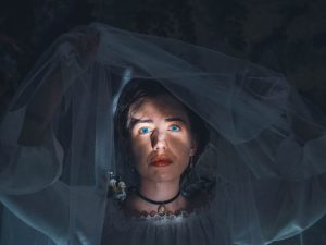 A dark photo in which a beam of light illuminates a woman's face, as she lifts a veil.