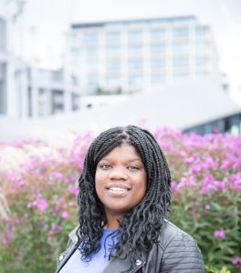 Anita is a black woman, with braid and she is looking and smiling at the camera.