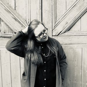 In a black and white photo, Annie is a white woman, with long hair. She wears glasses and is holding her arm up, looking away from the camera.