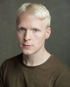 Lee Farrell, a man in his early 30s with platinum blonde hair, Blue eyes, wearing a dark green T-shirt with an intense, vulnerable expression to the camera. 