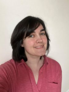 Louise is a white woman with dark mid-length hair with a side fringe wearing a pink blouse, smiling at the camera against a white background.