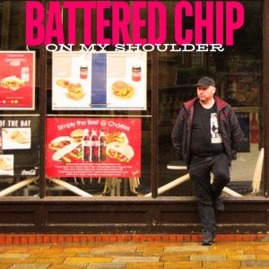 A man stands outside a fast food shop, looking away from the camera. Next to him are signs of burgers and fast food drinks. 