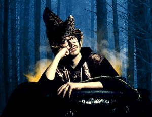 Edy, a man with black thick rimmed glasses and a moustache sits in a cauldron resting is head on his hand, wearing a wicker shaman hat and black robes, yellow smoke ascends from inside the cauldron.