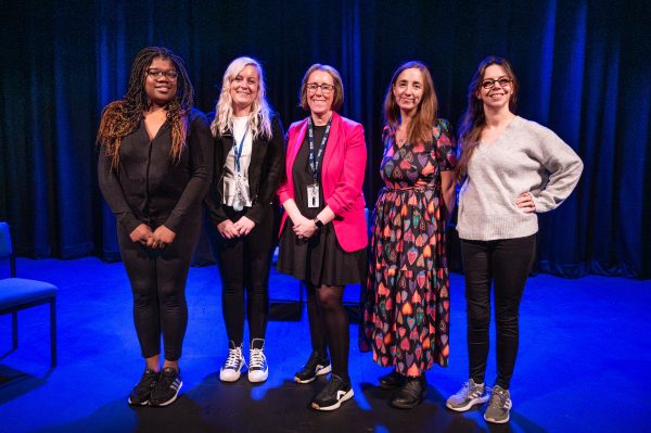 A group of 5 female panellists stand on a blue lit theatre stage.