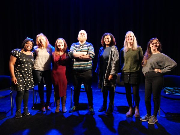 A group of 6 female panellists and a male panellist stand on a blue lit stage. 