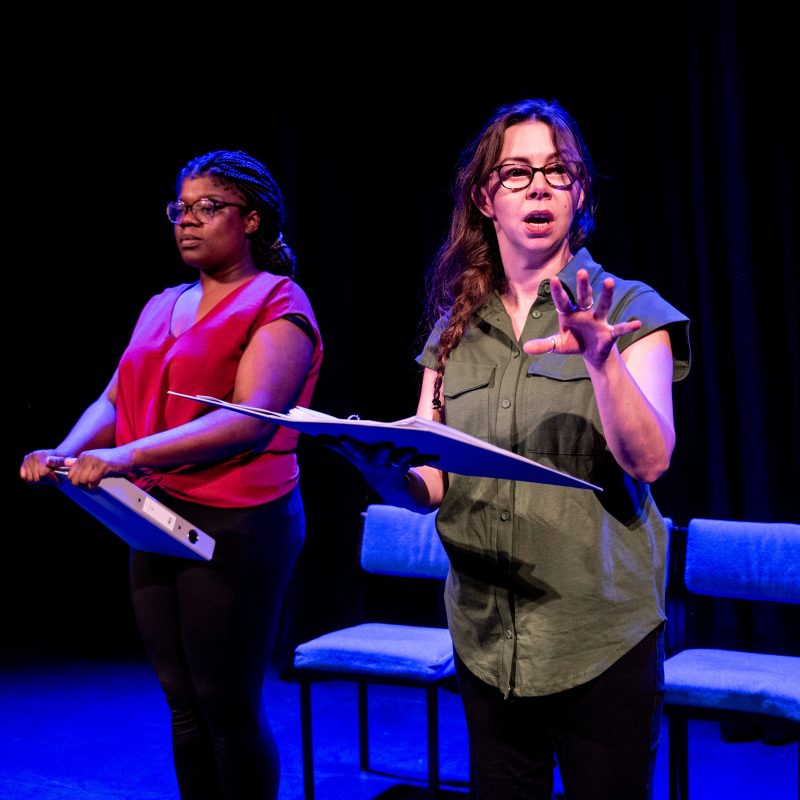 Two Actors stand on a stage in blue lighting. To the left a black woman holding a folder. She wears glasses and is dressed in a smart red top and black trousers. To the right a white woman holding a folder and speaking in mid sentence. She wears glasses and is dressed in a smart green top and black trousers.