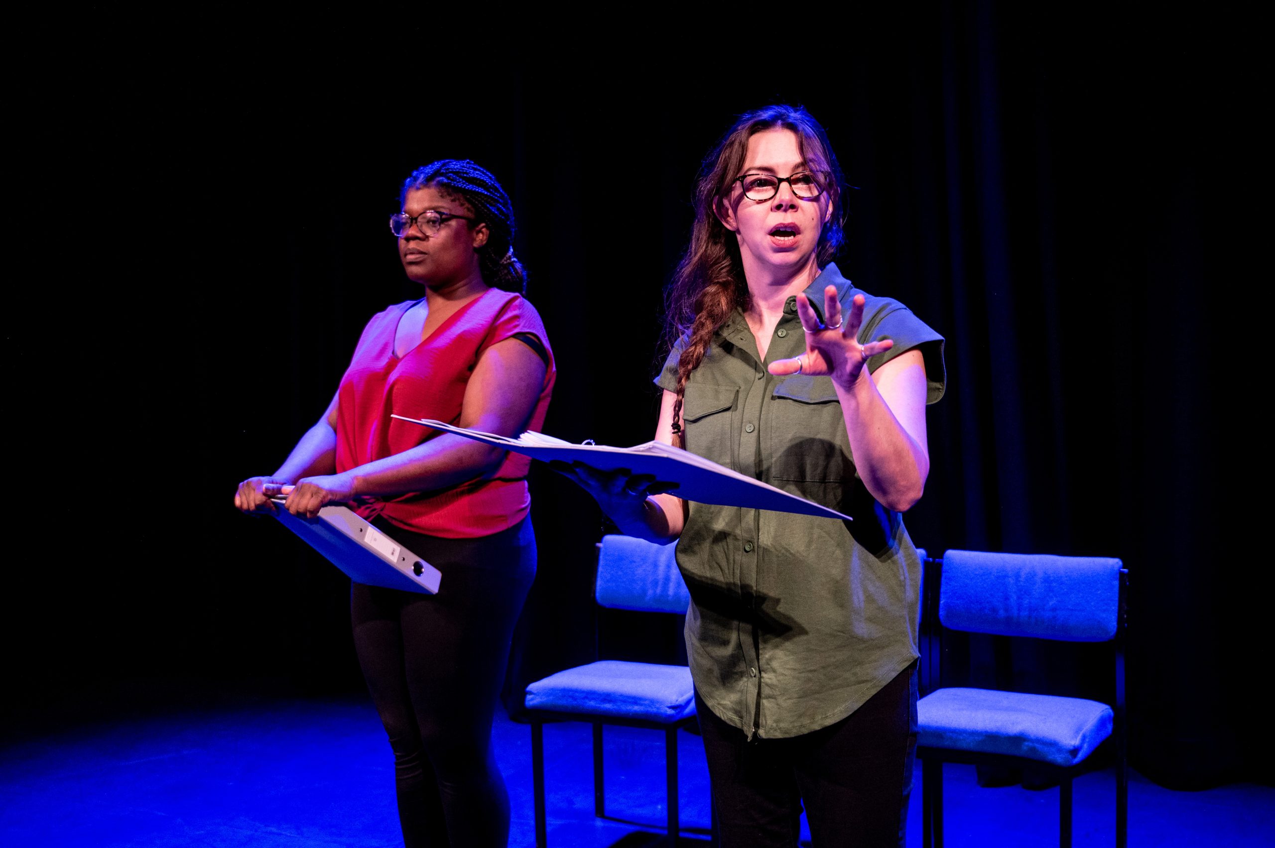 Two Actors stand on a stage in blue lighting. To the left a black woman holding a folder. She wears glasses and is dressed in a smart red top and black trousers. To the right a white woman holding a folder and speaking in mid sentence. She wears glasses and is dressed in a smart green top and black trousers.