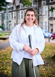 Tafsila is a South Asian female in her 40s is standing smiling in a park area holding her long white cane. She’s wearing a white t-shirt with a white open shirt and khaki green trousers.